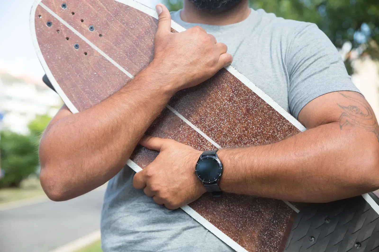 A man in a grey shirt outdoors holding a skateboard, wearing a wristwatch.